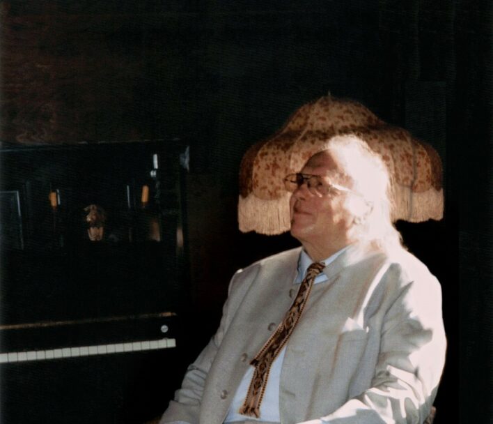 Edgars Račevskis sēž Jāņa Akuratera muzeja kamīnzālē, aiz viņa redzamas klavieres un lampa. Viss apkārt ir tumšs, diriģents tērpies gaišā uzvalkā. No loga nāk saules gaisma.