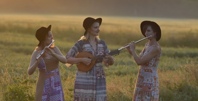 Grupas "Svīre" fotogrāfija. Trīs meitenes saulrietā stāv pļavā. Divas no meitenēm spēlē flautu, viena - mandolīnu.
