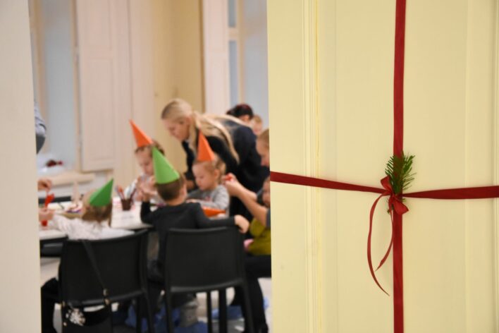 Foto priekšplānā redzamas baltas durvis, kurām apkārt apsieta sarkana lentīte un iesiets skuju zariņš. Pa pusatvērtajām durvīm fotogrāfijas tālākajā plānā redzami bērni, kuri darbojas ar papīru pie galda. Bērniem galvās zaļas un oranžas papīra svētku cepures.
