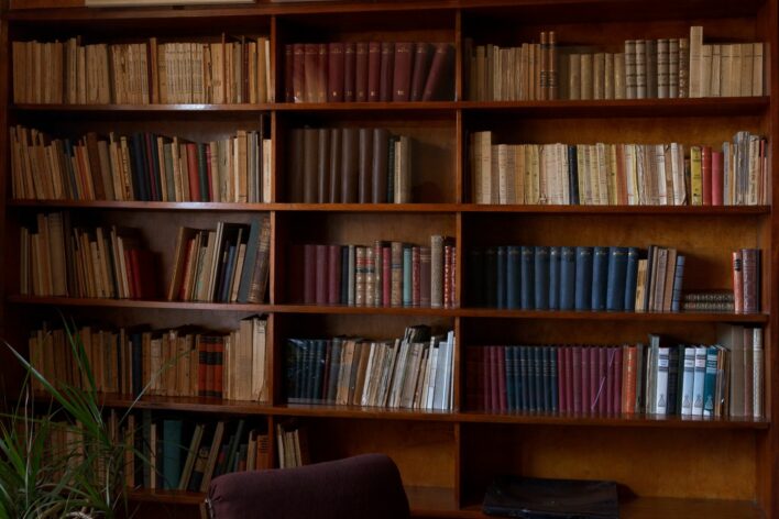 Grāmatplaukts Jāņa Akuratera kabinetā. Tas stiepjas gar visu sienu, plauktā redzamas grāmatu muguriņas, kas novietotas vertikāli