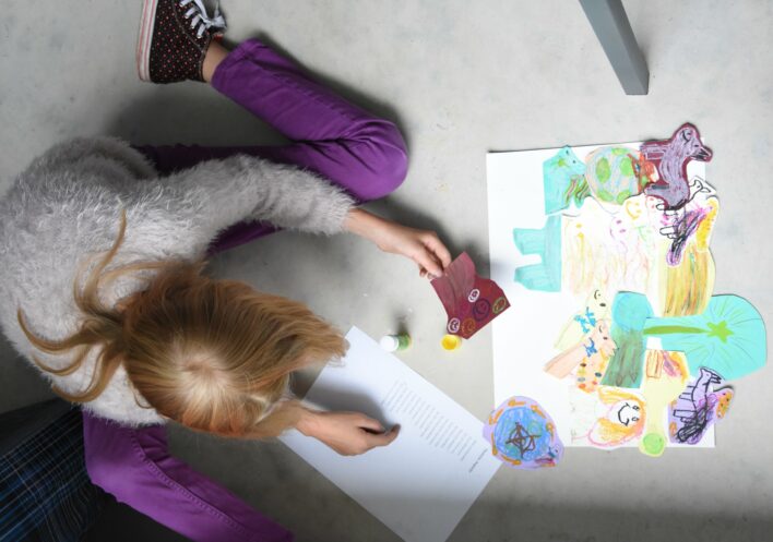 Meitene sēž uz grīdas un līmē kolāžu no pašas zīmētiem zīmējumiem.