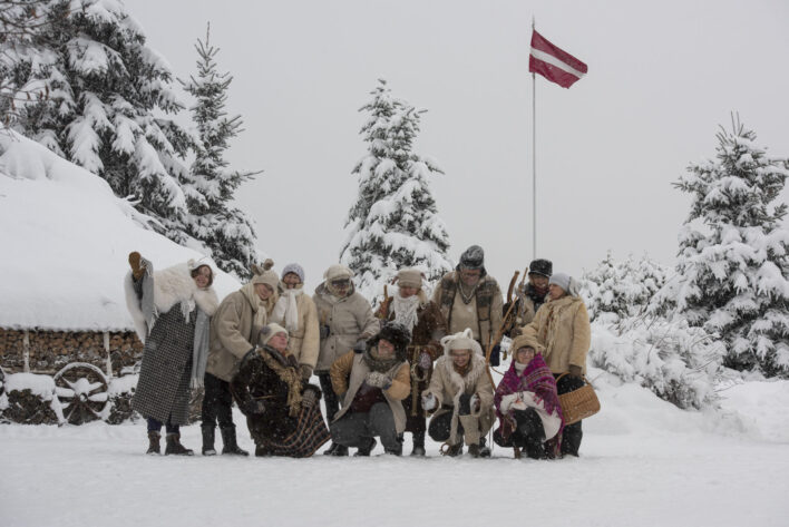 Tautas tradīciju kopa "Budēļi" ziemas dienā, visi saģērbušies maskās. Fonā apsnigušas egles, karogs. Foto: A. Eglītis