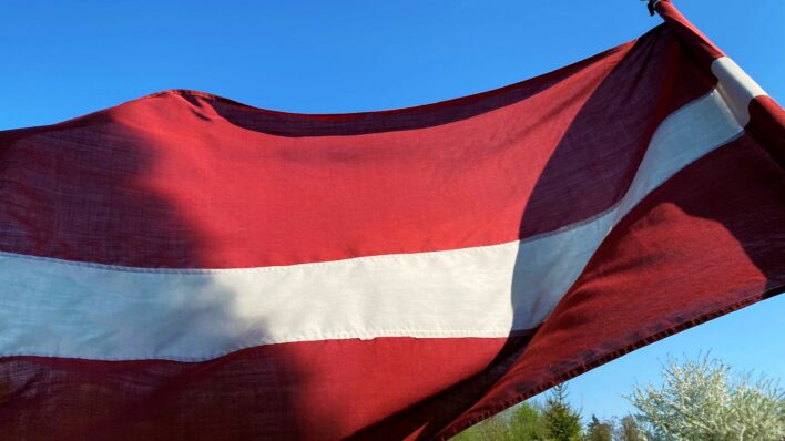 Plīvojošs Latvijas karogs, fonā redzamas zilas debesis bez mākoņiem