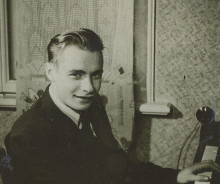 Ojārs Vācietis pie klavierēm, LVU Filoloģijas fakultātes 2. kursa students, 1954. gadā. Nezināms fotogrāfs. Ojārs sēž pie klavierēm, rokas uzlicis uz taustiņiem, skatās un fotogrāfu. Fonā ir logs ar aizkaru.