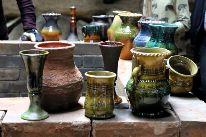 Keramikas trauki dažādās formās un krāsās izkārtoti uz ķieģeļu virsmas tradicionālā darbnīcā; fonā redzami cilvēki, kas vēro vai piedalās procesā.