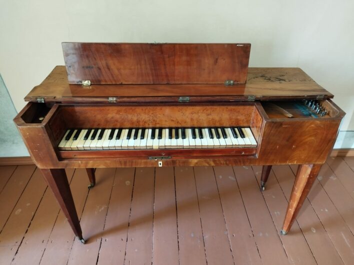 Koka korpusa tāfelklavieres ar atvērtu vāku, kas atsedz klaviatūru un stīgu mehānismu. Instruments stāv uz četrām slaidām kājām brūni krāsotā koka grīdas telpā. Klaviatūrai ir balti un melni taustiņi, un korpusa virsmā redzamas laika gaitā radušās nolietojuma pēdas.