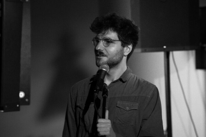 Melnbalts attēls, kurā redzams vīrietis ar brillēm un īsu bārdu, stāvot pie mikrofona un runājot uz skatuves. Viņš ir ģērbies pogājamā kreklā un skatās sānis, fonā redzama telpas iekārtojuma detaļas.