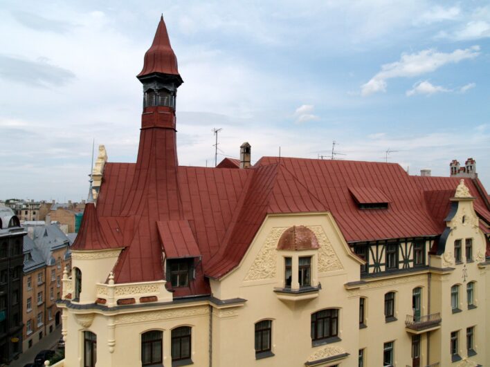 Skats uz jūgendstila ēku ar gaišu fasādi, dekoratīviem ciļņiem, sarkanu jumtu un augstu torni. Fonā redzamas Rīgas centra ēkas un zilas, daļēji mākoņainas debesis.